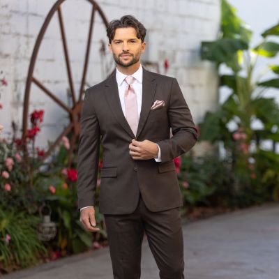 Man in a brown suit with white shirt and pink tie and pocket square standing outdoors on a patio