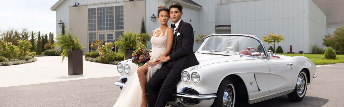 man in black tuxedo sitting next to bride in white dress with slit on a classic car in front of the reception venue