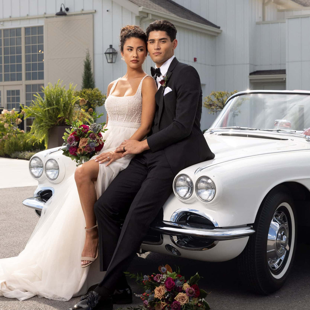 man in black tuxedo sitting next to bride in white dress with slit on a classic car with fall colored flowers next to them
