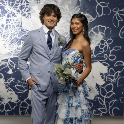 Man in oxford blue suit with a navy blue windsor tie standing next to a girl in a blue and white floral, ruffled dress. 